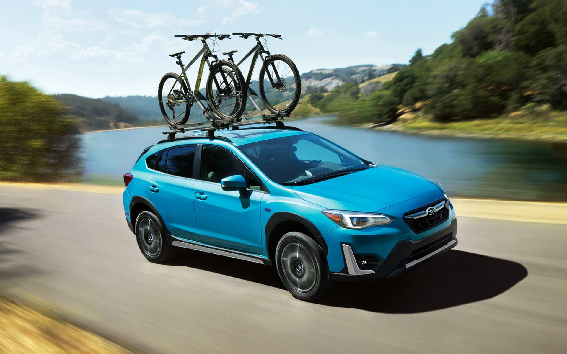 A blue Crosstrek Hybrid with two bicycles on its roof rack driving beside a river | River City Subaru in Huntington WV